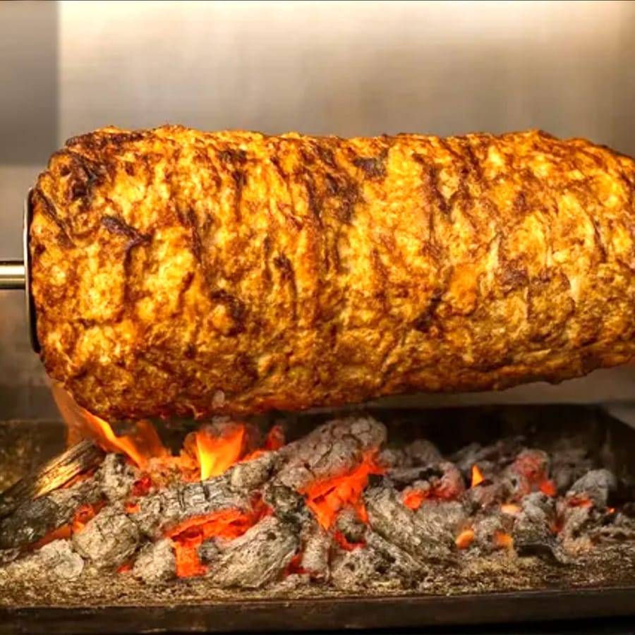 A large meat roll on a rotisserie spit above glowing charcoal embers with open flame, a bowl of white sauce and a bowl of torshi in the foreground.